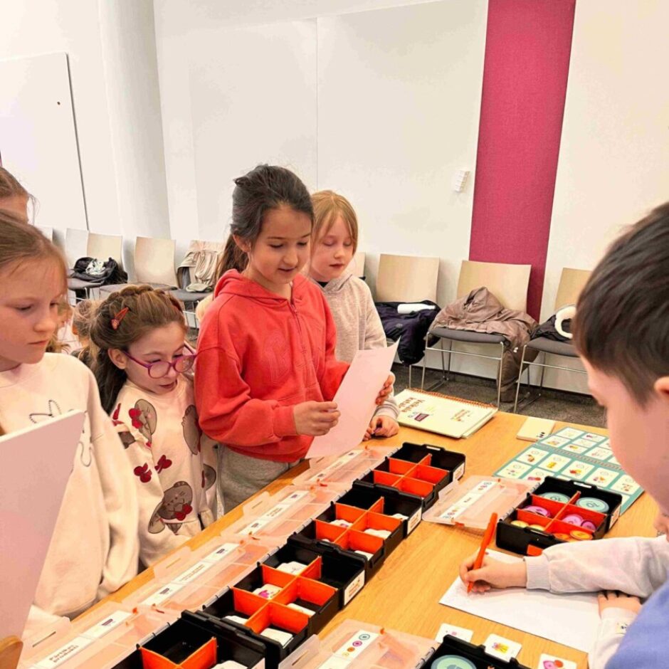 Bērnu grupa pie galda spēlē izglītojošu spēli ar kartītēm un žetoniem. Meitene sarkanā džemperī lasa rozā papīra lapu, kamēr cits bērns raksta uz baltas lapas. Uz galda ir melni konteineri ar baltām kartītēm, kas attēlo ikonas, un krāsainiem apaļiem žetoniem ar skaitļiem, piemēram, 5, 10 un 40.