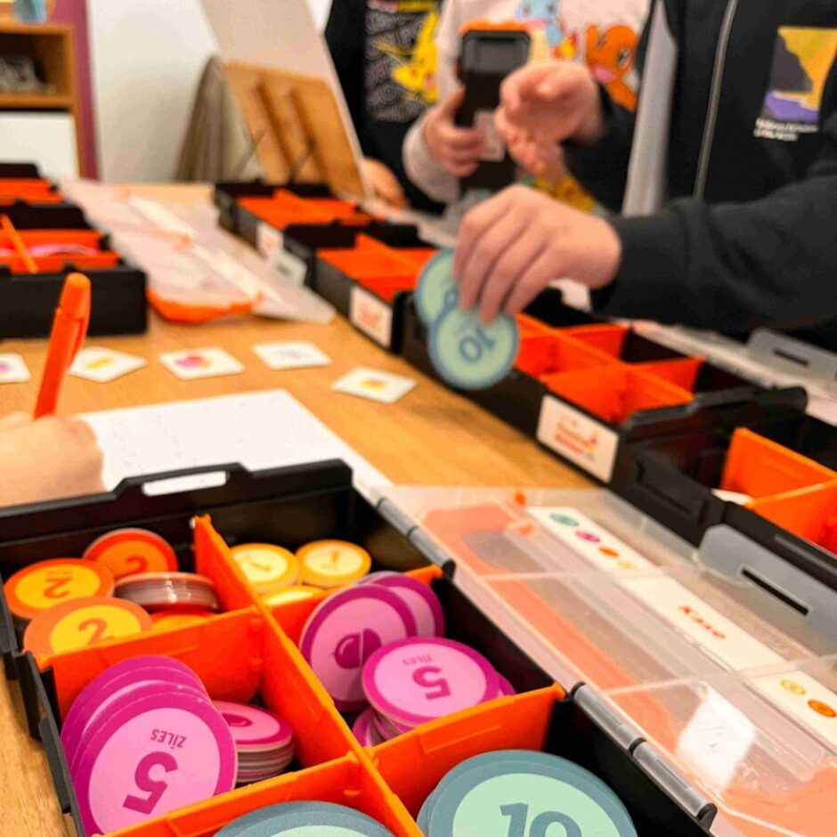 Uz koka galda redzamas melnas paplātes ar oranžiem nodalījumiem, pilnas ar krāsainiem žetoniem. Priekšplānā skaidri redzami zili-zaļi žetoni ar tekstu 