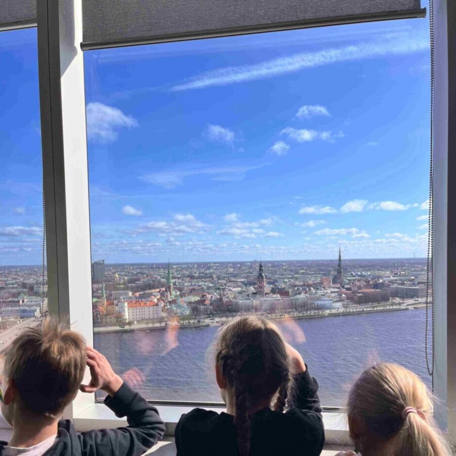 Trīs bērni no mugurpuses skatās pa lielu logu uz Rīgas panorāmu ar Daugavu, pilsētas ēkām un baznīcu torņiem zem zilām, mākoņainām debesīm.