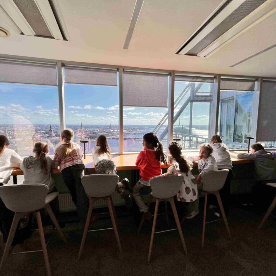 Grupa bērnu no mugurpuses sēž pie gara galda lielā loga priekšā, vērojot plašu panorāmas skatu uz pilsētu ar upi, tiltiem un baznīcu torņiem saulainā dienā.