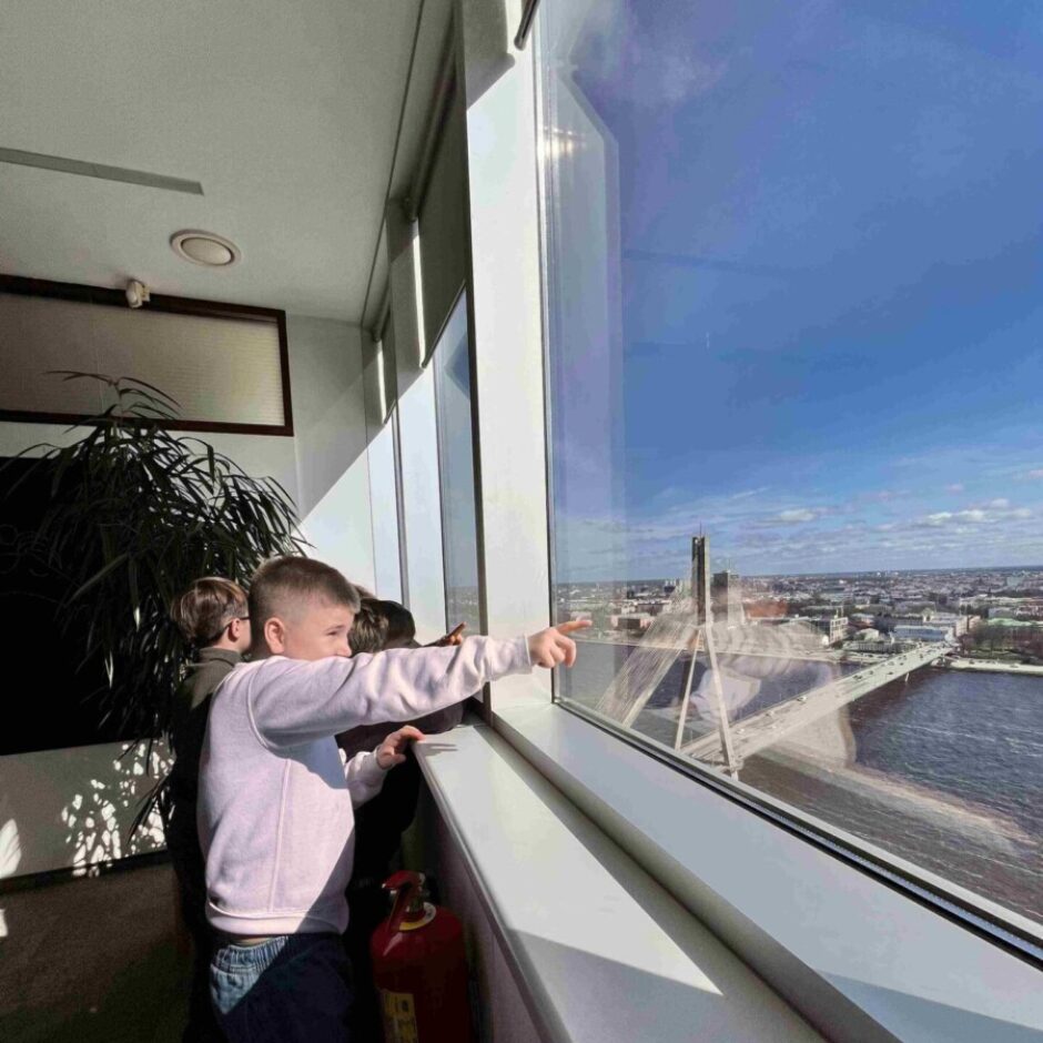 Zēns gaišā džemperī rāda ar pirkstu pa plašu logu uz Rīgas pilsētas panorāmu, kur redzams Vanšu tilts pāri Daugavai. Vairāki bērni skatās ārā saulainā dienā.