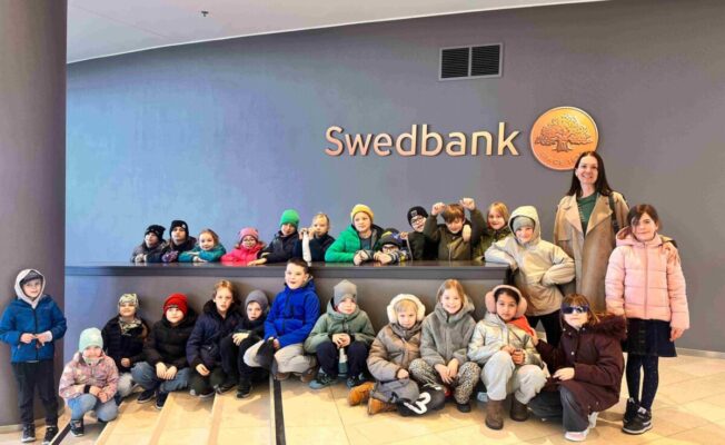 Grupa bērnu un pieauguša sieviete pozē fotogrāfijai pie sienas ar uzrakstu 