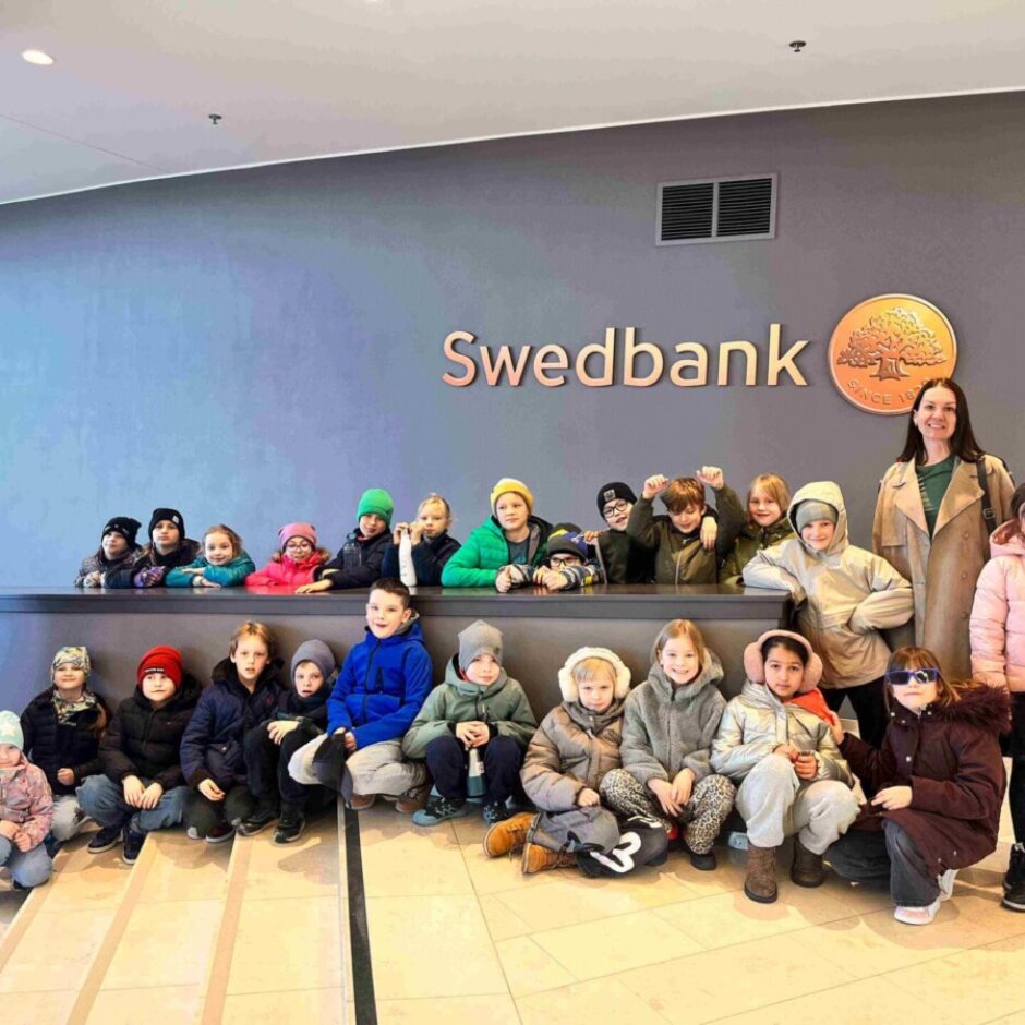 Grupa bērnu un pieauguša sieviete pozē fotogrāfijai pie sienas ar uzrakstu 