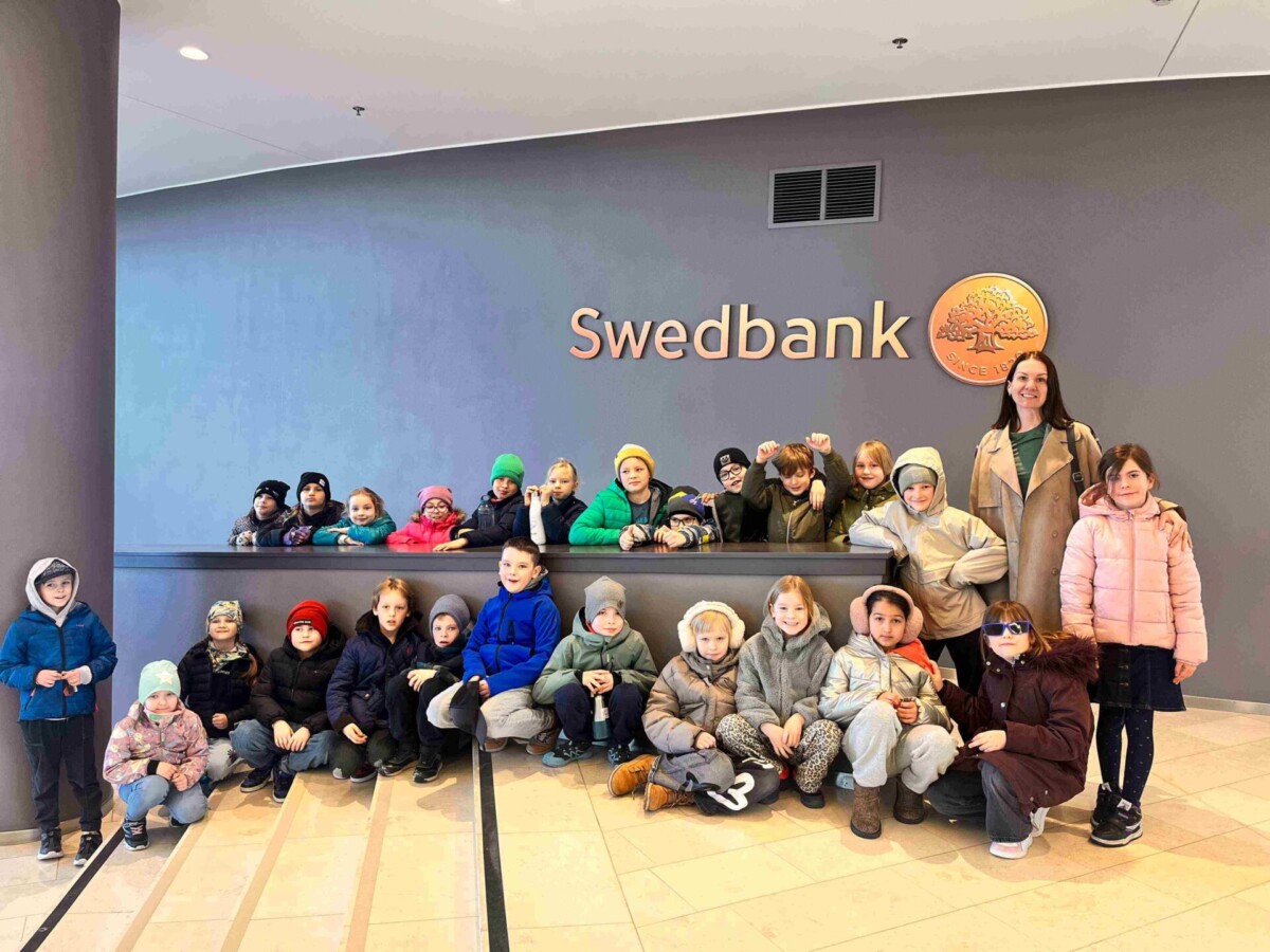 Grupa bērnu un pieauguša sieviete pozē fotogrāfijai pie sienas ar uzrakstu 