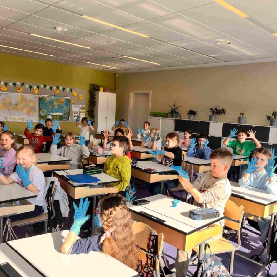 Skolēni sēž gaišā klasē, valkājot gaiši zilus cimdus un priecīgi paceļot rokas. Fonā redzamas kartes un citi mācību plakāti.