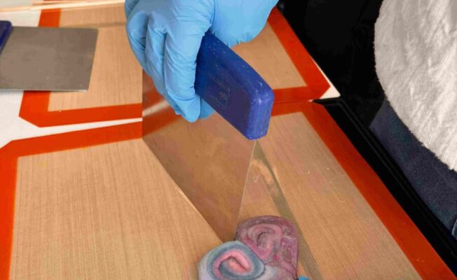 Zilajos cimdos tērpta roka ar instrumentu veido sirds formas, virpuļveida rozā, violetu un zilu konfekti uz silikona paklājiņa. Blakus redzami koka kociņi popu izgatavošanai.