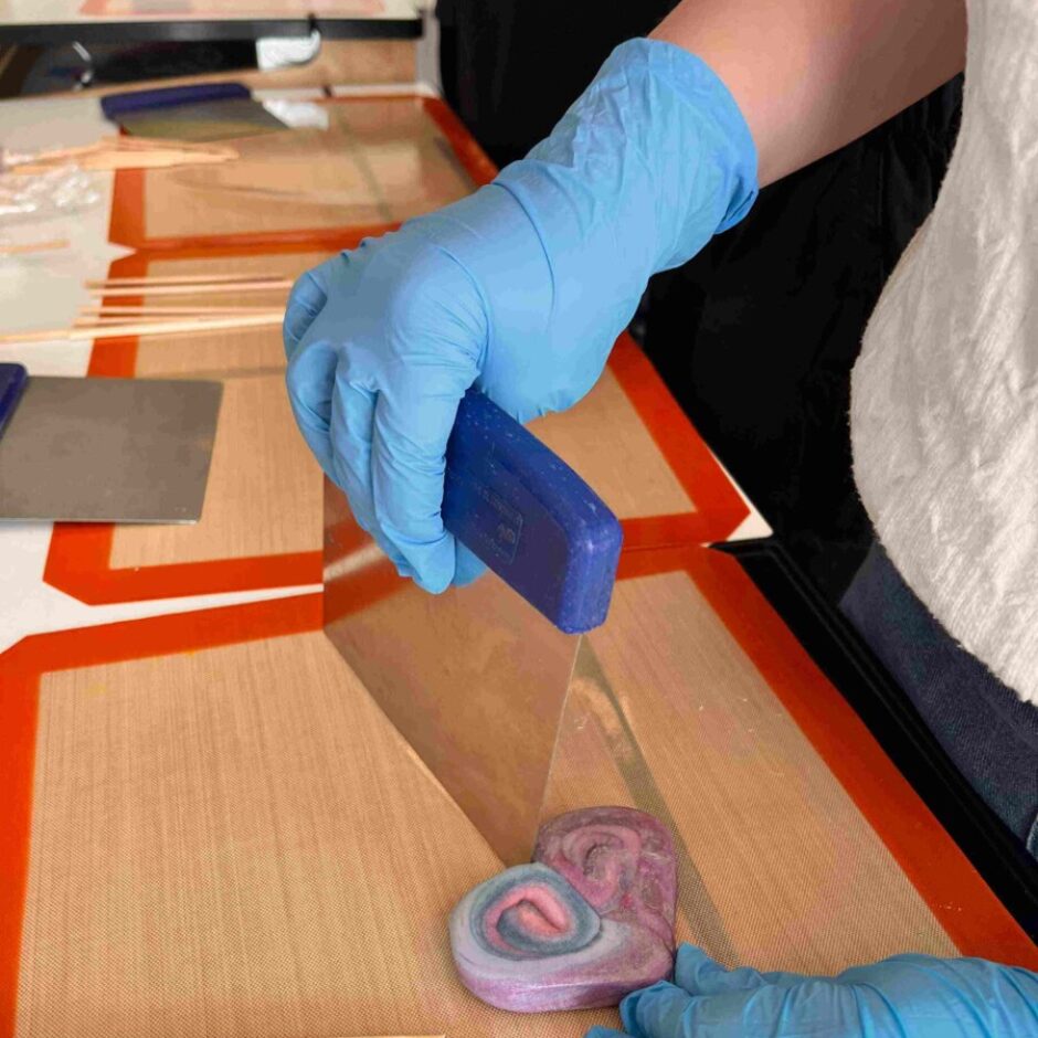 Zilajos cimdos tērpta roka ar instrumentu veido sirds formas, virpuļveida rozā, violetu un zilu konfekti uz silikona paklājiņa. Blakus redzami koka kociņi popu izgatavošanai.