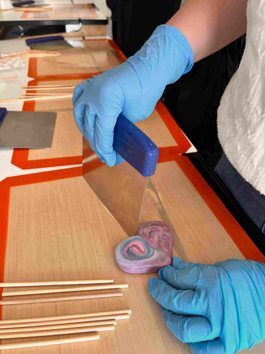 Zilajos cimdos tērpta roka ar instrumentu veido sirds formas, virpuļveida rozā, violetu un zilu konfekti uz silikona paklājiņa. Blakus redzami koka kociņi popu izgatavošanai.