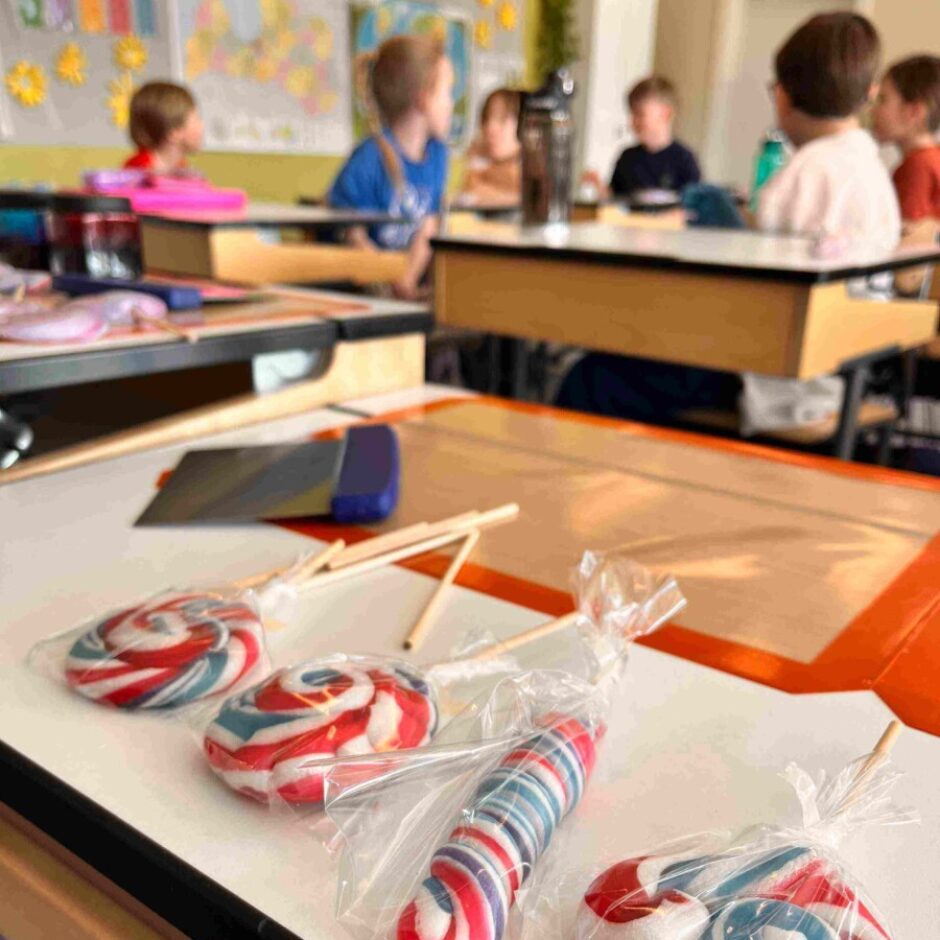 Četras sarkanbaltzilas konfektes-īkšķi plastmasas iesaiņojumā, dažādās formās (spirālveida, cilindriskas, izliektas), novietotas uz galda. Aizmugurē redzama izplūdusi klases telpa ar bērniem, kas sēž pie galdiem.