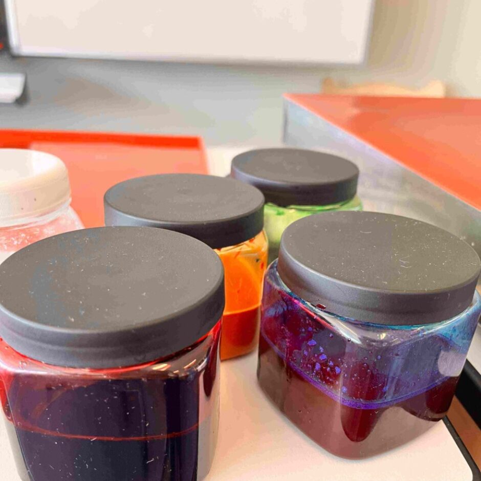 Vairākas caurspīdīgas burciņas ar melniem vāciņiem, kurās ir spilgti krāsaini šķidrumi vai geli – tumši sarkans, oranžs, zaļš un sarkans ar zilu mirdzumu, novietotas uz balta paliktņa.