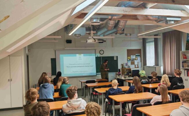 Gaiša telpa ar atklātām koka sijām, kurā studenti sēž pie koka galdiem un klausās sievietes prezentāciju. Sieviete stāv pie projekcijas ekrāna, uz kura redzams teksts: 
