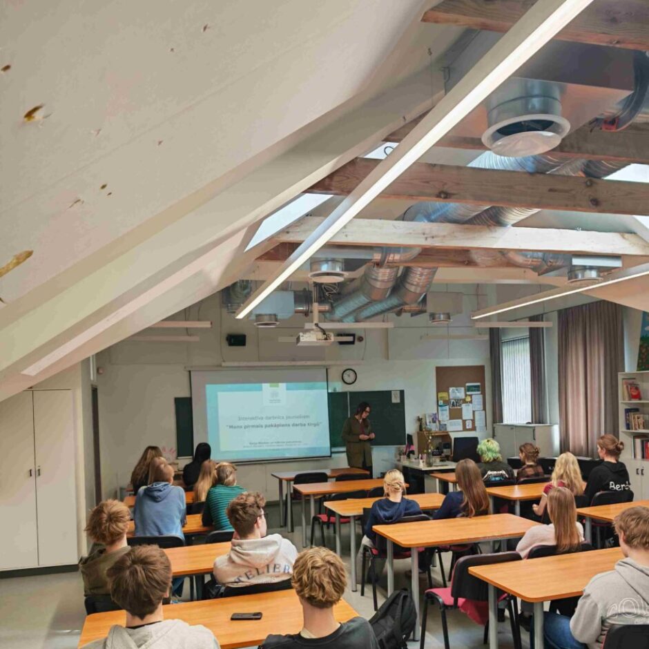 Gaiša telpa ar atklātām koka sijām, kurā studenti sēž pie koka galdiem un klausās sievietes prezentāciju. Sieviete stāv pie projekcijas ekrāna, uz kura redzams teksts: 