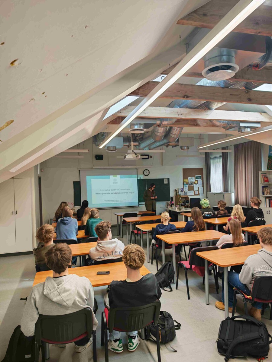 Gaiša telpa ar atklātām koka sijām, kurā studenti sēž pie koka galdiem un klausās sievietes prezentāciju. Sieviete stāv pie projekcijas ekrāna, uz kura redzams teksts: 