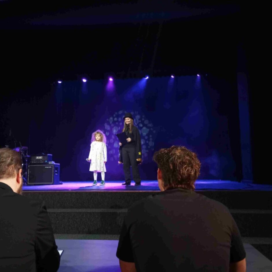 Uz skatuves stāv jauna meitene ar gariem, gaišiem matiem baltā kleitā un sieviete melnā apģērbā, kura rokās tur planšeti vai kladi. Fona ekrānā redzami balti, animēti spoki. Divi vīrieši priekšplānā vēro notiekošo no mugurpuses.