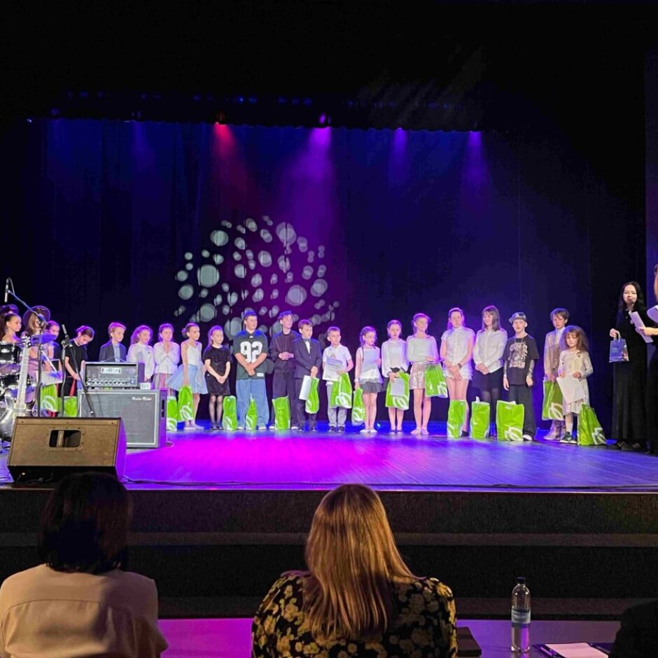 Bērnu grupa stāv uz tumšas skatuves, ko izgaismo purpura un zilas gaismas. Daudzi tur rokās sertifikātus, pie kājām stāvot zaļiem dāvanu maisiņiem. Skatuves labajā pusē divi pieaugušie pasniedz balvas, viena sieviete tērpā ar zvaigznēm. Kreisajā pusē redzams bungu komplekts un mūzikas aprīkojums. Viens zēns centrā valkā zilu kreklu ar numuru 