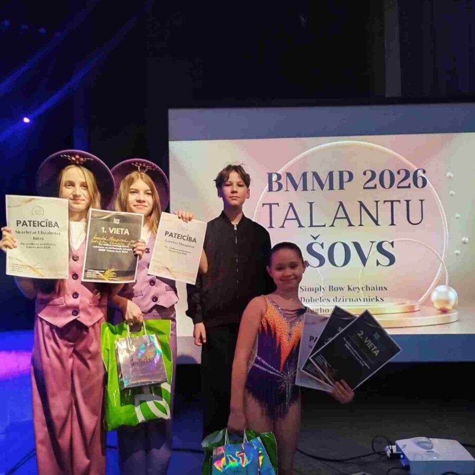 Četri bērni – trīs meitenes un viens puika – stāv uz skatuves, turot sertifikātus un dāvanu maisiņus. Divas meitenes kreisajā pusē purpura tērpos ar platmalēm tur sertifikātus ar uzrakstiem 