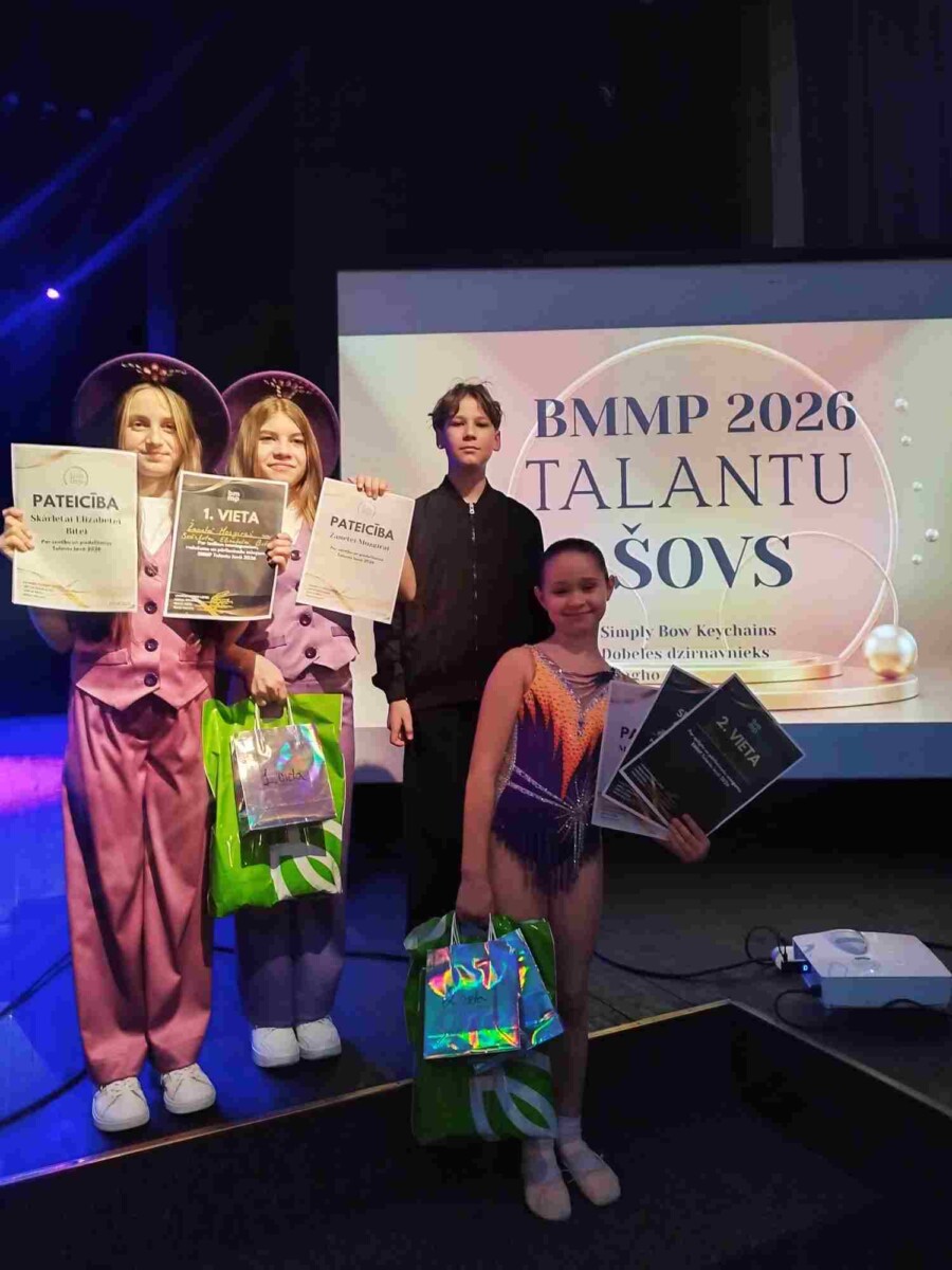 Četri bērni – trīs meitenes un viens puika – stāv uz skatuves, turot sertifikātus un dāvanu maisiņus. Divas meitenes kreisajā pusē purpura tērpos ar platmalēm tur sertifikātus ar uzrakstiem 