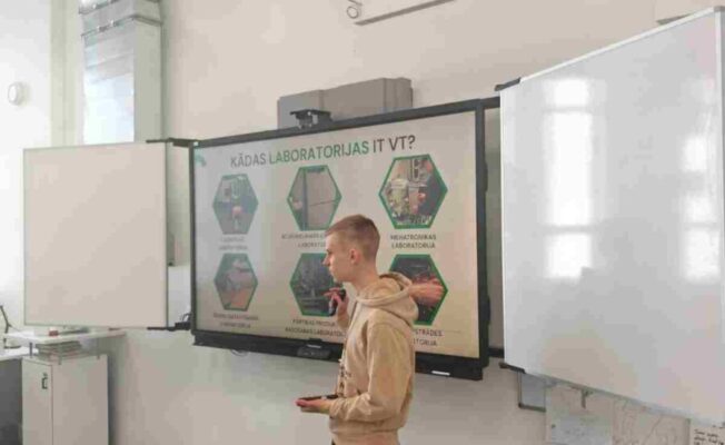 Jauns vīrietis gaišā džemperī stāv klases telpā pie liela interaktīvā ekrāna, uz kura redzama prezentācija ar virsrakstu 