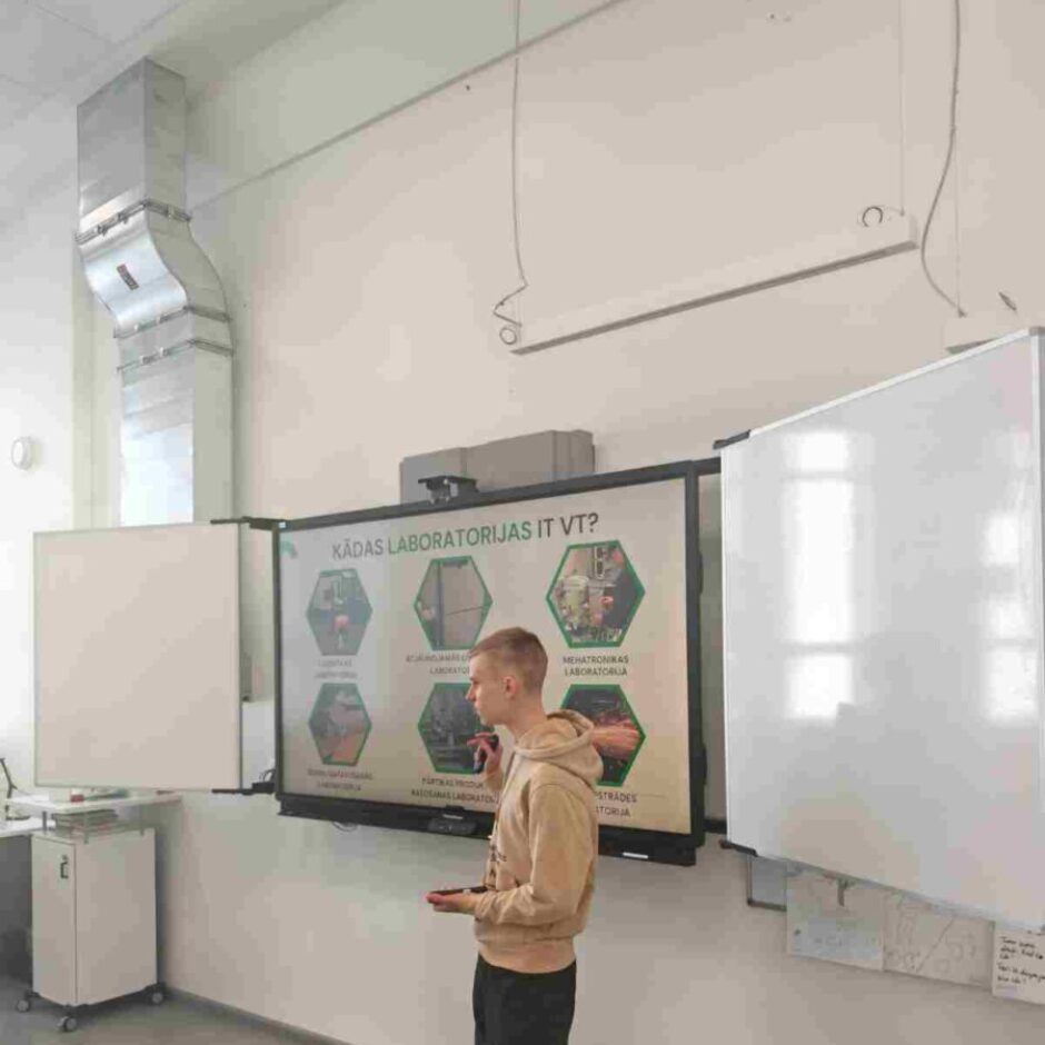 Jauns vīrietis gaišā džemperī stāv klases telpā pie liela interaktīvā ekrāna, uz kura redzama prezentācija ar virsrakstu 