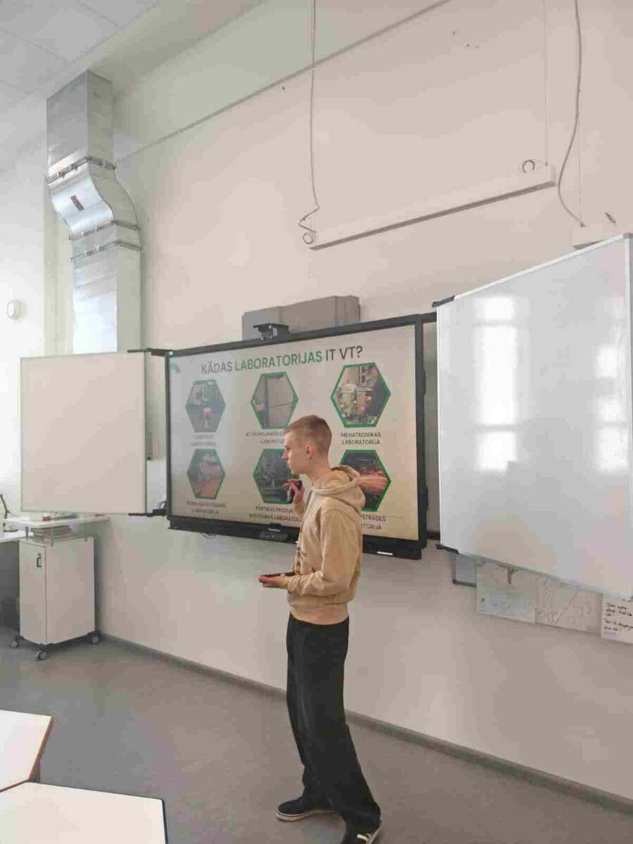 Jauns vīrietis gaišā džemperī stāv klases telpā pie liela interaktīvā ekrāna, uz kura redzama prezentācija ar virsrakstu 