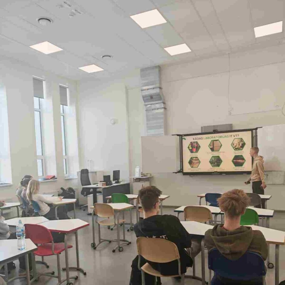 Skolēni sēž klasē, klausoties skolotāja prezentāciju uz liela ekrāna. Uz ekrāna redzams jautājums 