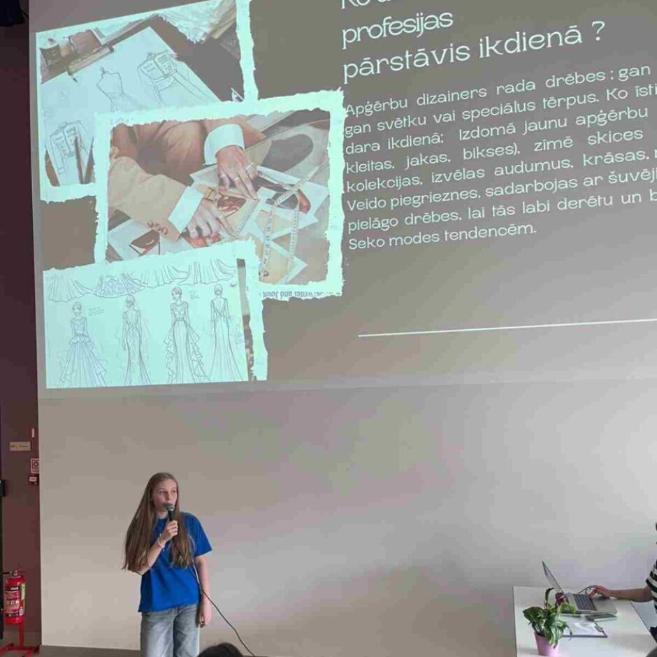 Jauna meitene stāv telpā ar mikrofonu rokā, uzstājoties auditorijas priekšā. Aiz viņas uz lielā ekrāna redzams slaids ar tekstu 