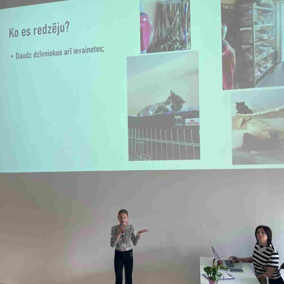 Jauna meitene stāv, turot mikrofonu un žestikulējot, pie projekcijas ekrāna, kurā redzams teksts 