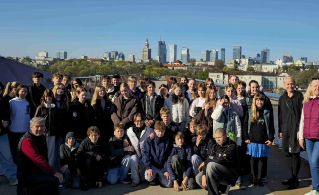 Liela skolēnu un vairāku pieaugušo grupa pozē kopīgai fotogrāfijai saulainā dienā, ar Varšavas debesskrāpju siluetu fonā.