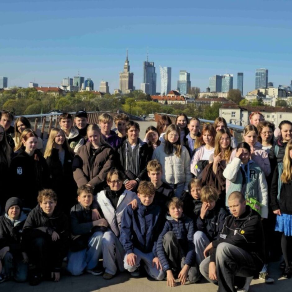 Liela skolēnu un vairāku pieaugušo grupa pozē kopīgai fotogrāfijai saulainā dienā, ar Varšavas debesskrāpju siluetu fonā.