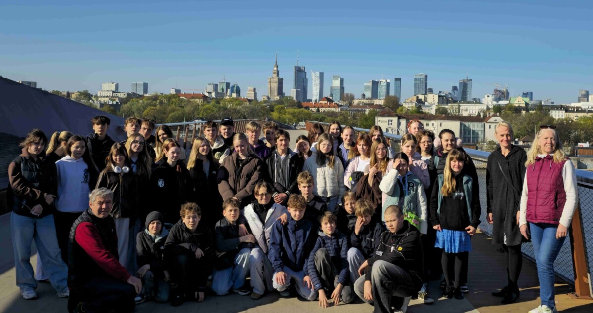 Liela skolēnu un vairāku pieaugušo grupa pozē kopīgai fotogrāfijai saulainā dienā, ar Varšavas debesskrāpju siluetu fonā.