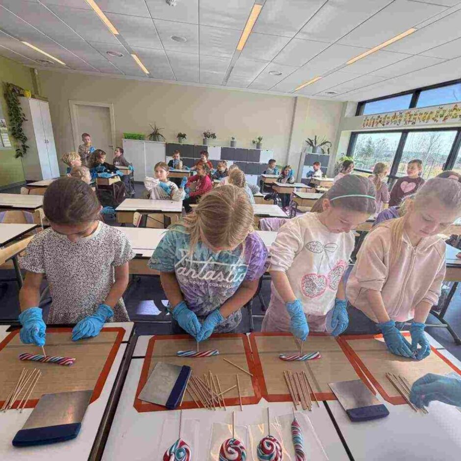 Klasē bērni zilajos cimdos formē svītrainas konfektes uz silikona paklājiņiem. Priekšplānā redzamas gatavas konfektes uz kociņa, fonā sēž citi skolēni, un labajā pusē redzama daļa pieaugušā.