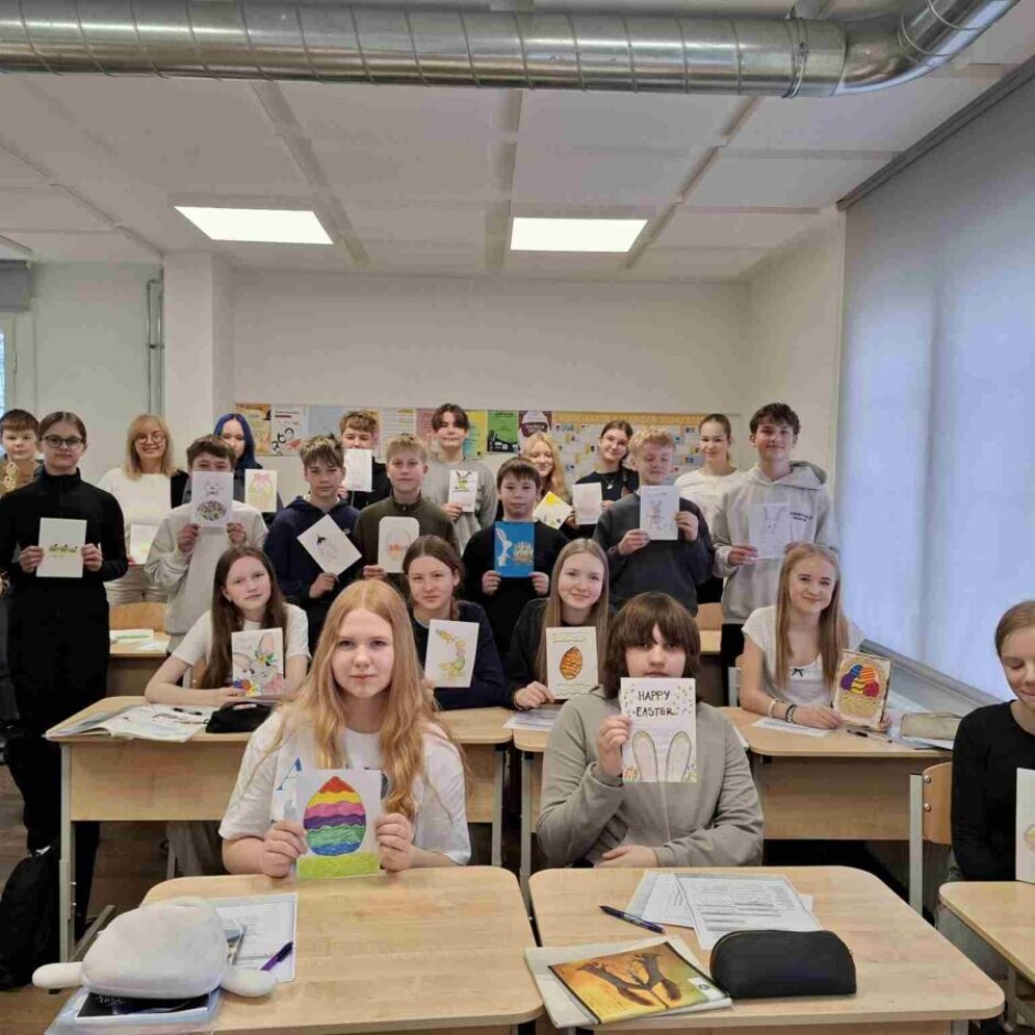 Apmēram divdesmit skolēnu un skolotāja pozē klasē, turot rokās Lieldienu tematiskus zīmējumus vai apsveikuma kartītes ar zaķiem un krāsainām olām. Viena kartīte vēsta 