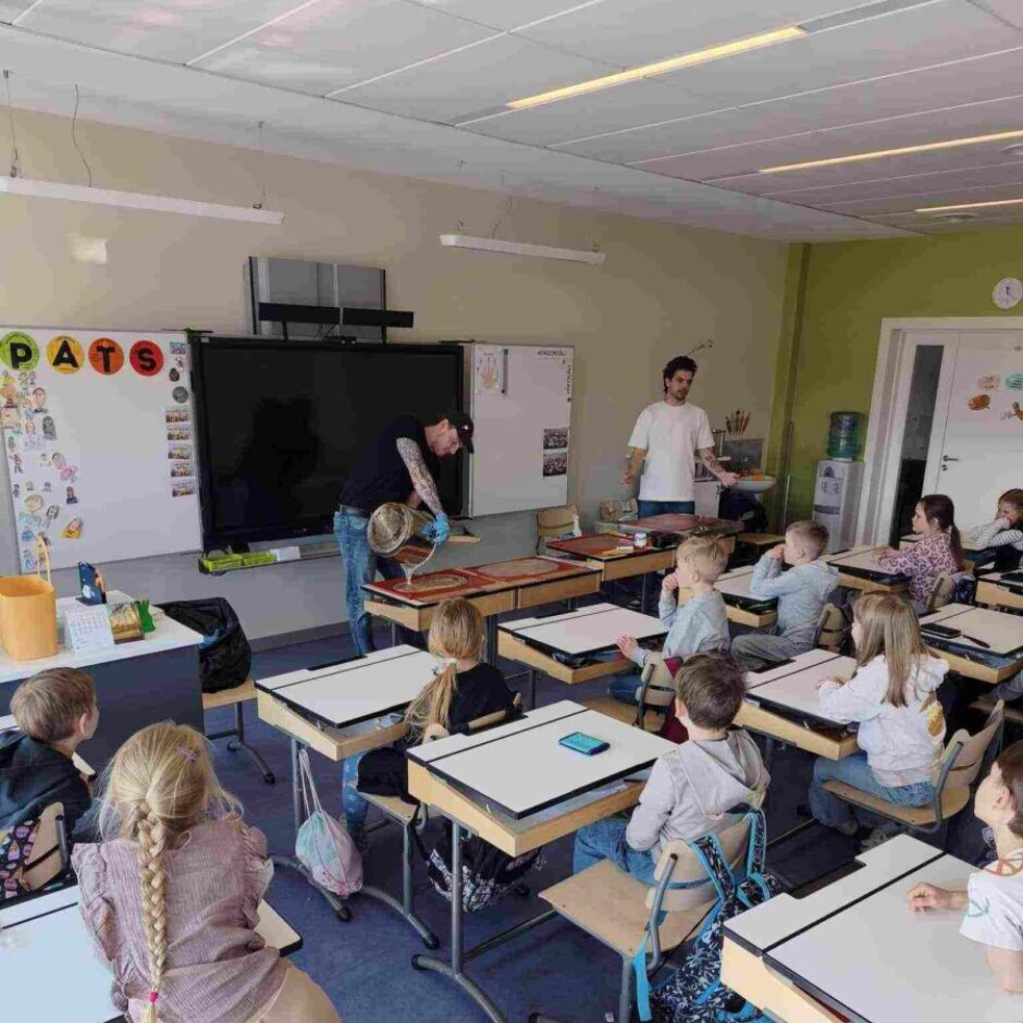 Klase, kurā divi pieaugušie demonstrē eksperimentu vai rokdarbus bērniem. Vīrietis tumšā kreklā un zilajos cimdos lej šķidrumu no metāla trauka uz galda, kamēr otrs vīrietis baltā kreklā stāv blakus. Bērni uzmanīgi sēž pie soliem un vēro. Uz baltās tāfeles redzami krāsaini burti 