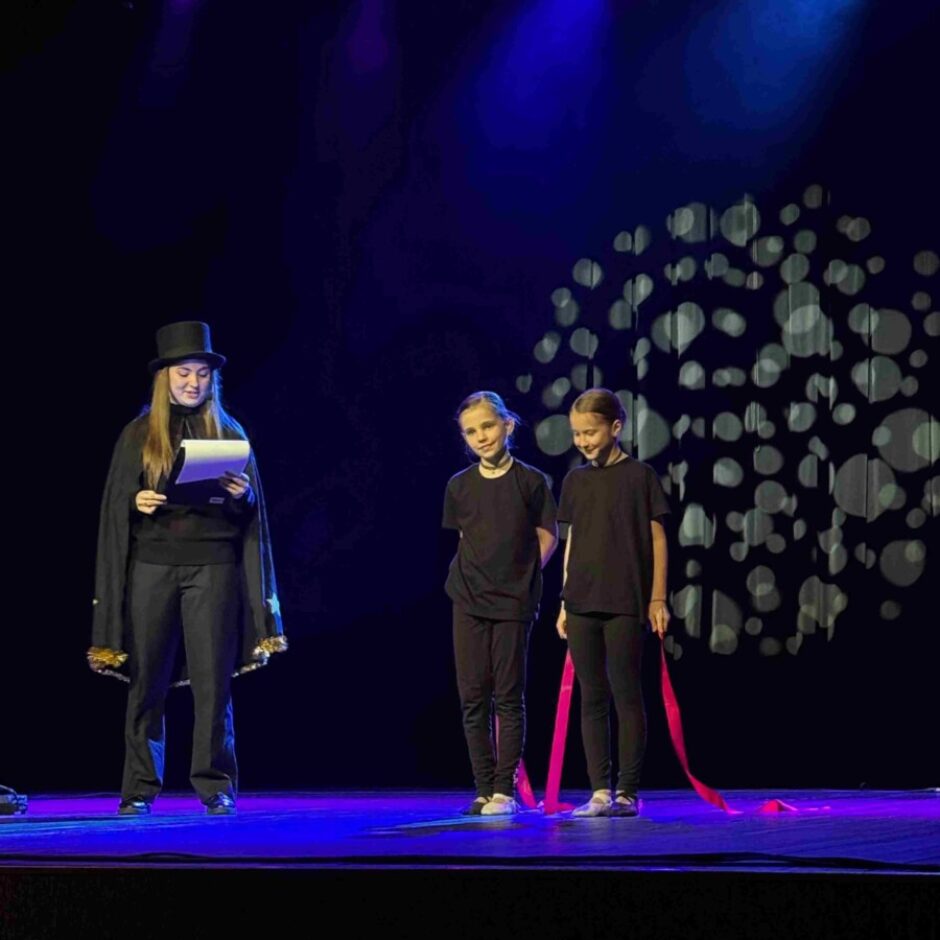 Trīs cilvēki uz tumšas skatuves. Kreisajā pusē jauna persona melnā cilindrā un apmetnī lasa no papīra. Labajā pusē stāv divas jaunas meitenes melnos kreklos un biksēs, viena no tām tur sarkanu lentu. Skatuve ir izgaismota zilām gaismām, un fonā ir gaišu apļu raksts.