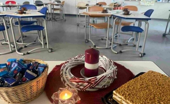 Priekšplānā galds ar pītu grozu, pilnu ar šokolādes konfektēm, sarkanbaltsarkanu sveci un vainagu, iedegtu tējas sveci un kūku ar drupačām. Fonā redzama klase ar vairākiem galdiem un krēsliem, uz kuriem stāv krūzes.