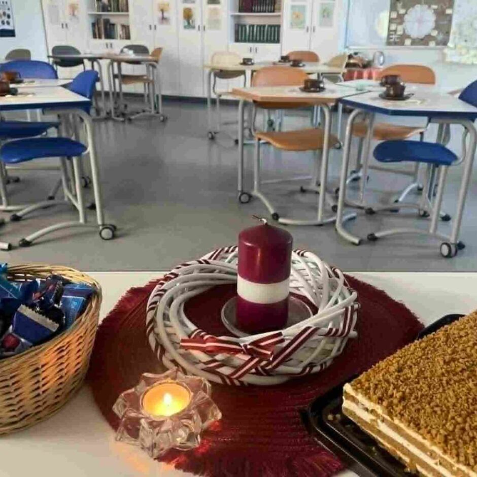 Priekšplānā galds ar pītu grozu, pilnu ar šokolādes konfektēm, sarkanbaltsarkanu sveci un vainagu, iedegtu tējas sveci un kūku ar drupačām. Fonā redzama klase ar vairākiem galdiem un krēsliem, uz kuriem stāv krūzes.