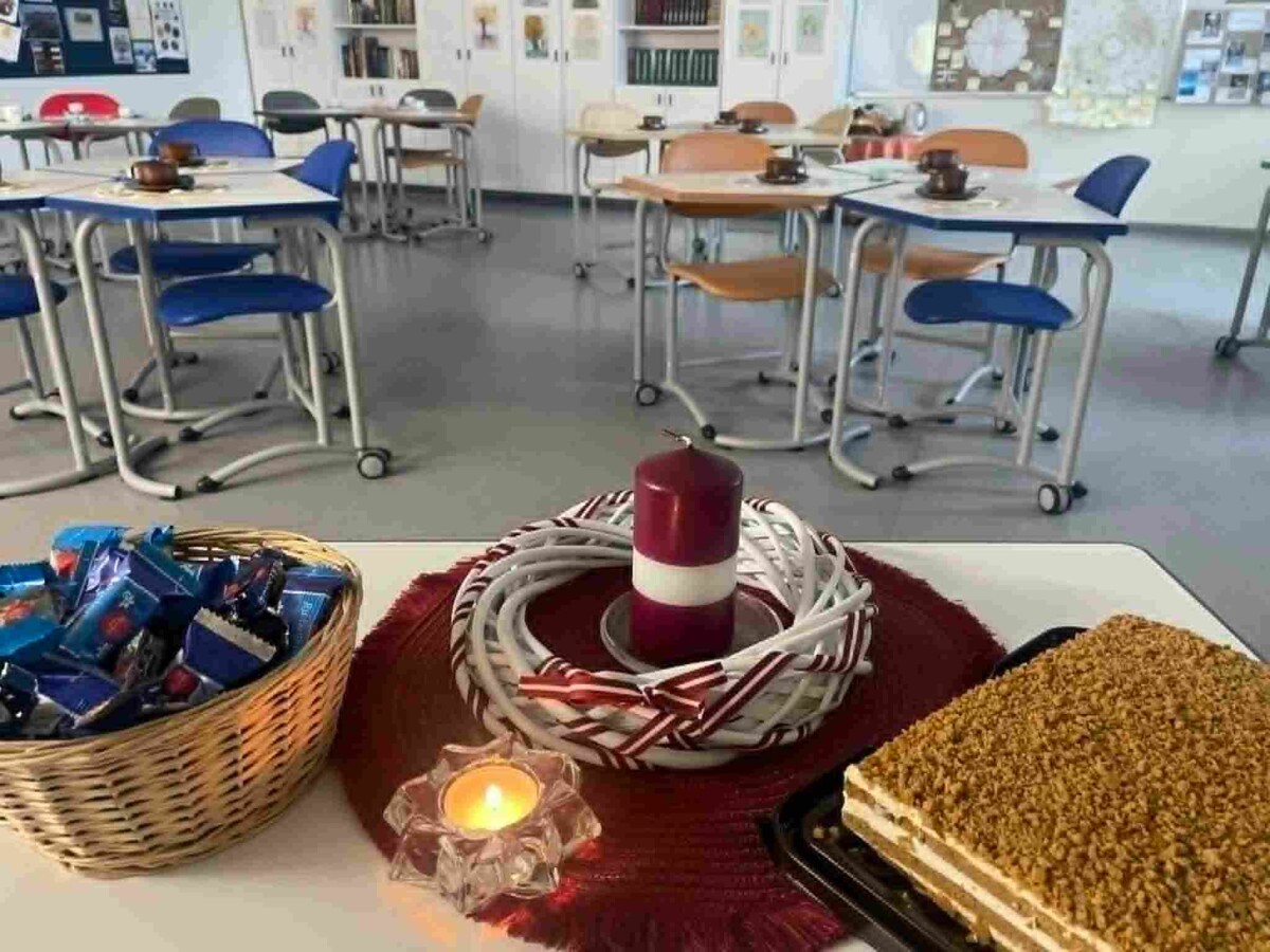Priekšplānā galds ar pītu grozu, pilnu ar šokolādes konfektēm, sarkanbaltsarkanu sveci un vainagu, iedegtu tējas sveci un kūku ar drupačām. Fonā redzama klase ar vairākiem galdiem un krēsliem, uz kuriem stāv krūzes.