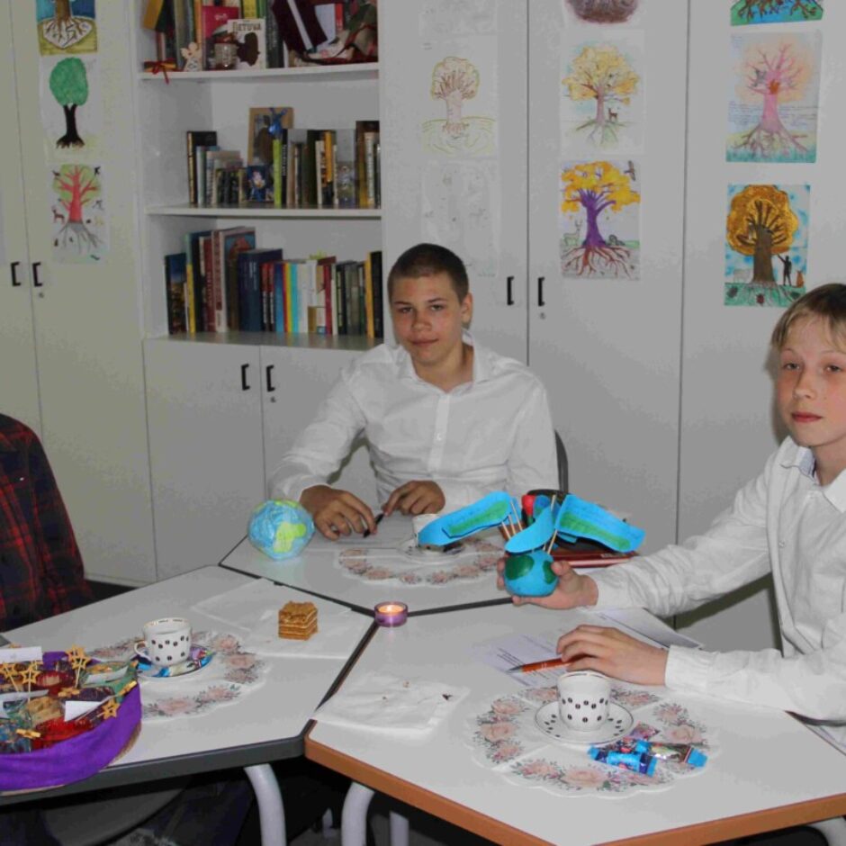 Trīs zēni sēž pie galdiem telpā, kas dekorēta ar bērnu zīmējumiem, kuros attēloti koki. Kreisajā pusē zēns rūtainā kreklā, viņam priekšā ir dekoratīvs projekts, kas atgādina laivu. Vidējais zēns baltā kreklā viegli smaida, viņam priekšā ir mazs zemeslodes modelis. Labajā pusē zēns baltā kreklā tur rokās zilu un zaļu modeli, kas piestiprināts pie nelielas zemeslodes.