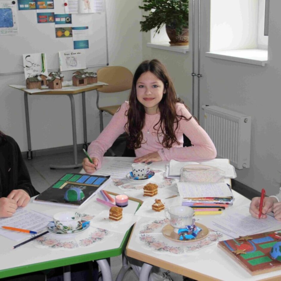 Trīs smaidīgas meitenes sēž pie galdiem klasē, ap kuriem izvietoti rakstāmpiederumi, rokdarbi, tējas krūzītes un globusa modelis. Divas meitenes raksta. Fonā redzamas tāfeles ar plakātiem un zīmējumiem.