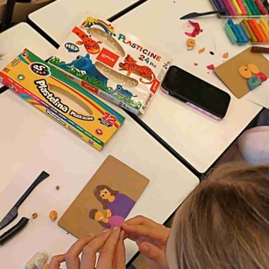 Bērns veido plastilīna figūriņas uz galda. Viņa rokas veido detaļas brūnai kartona pamatnei, uz kuras jau redzams attēls ar sievieti, kas tur bērnu. Uz galda ir arī plastilīna kastes ar uzrakstiem 
