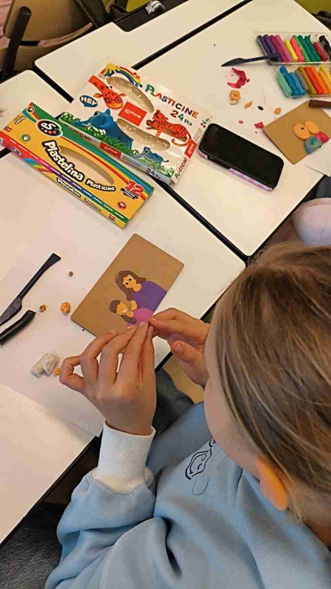 Bērns veido plastilīna figūriņas uz galda. Viņa rokas veido detaļas brūnai kartona pamatnei, uz kuras jau redzams attēls ar sievieti, kas tur bērnu. Uz galda ir arī plastilīna kastes ar uzrakstiem 