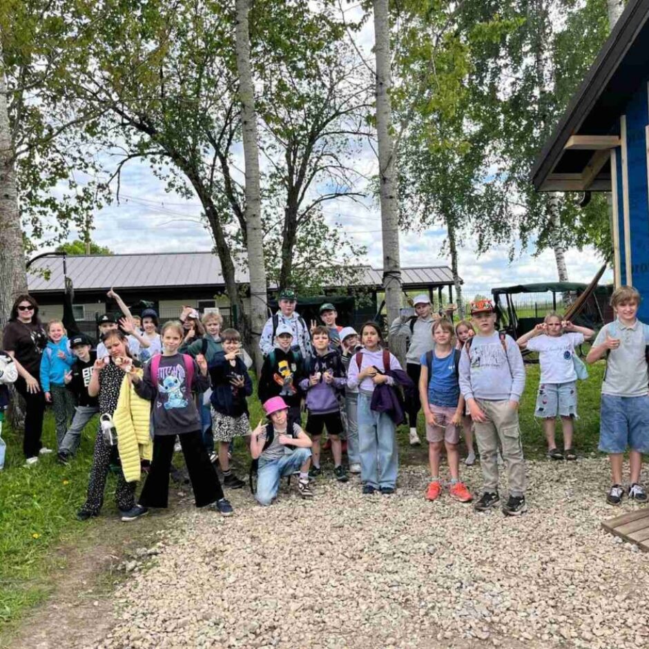 Liela bērnu grupa ar pieaugušajiem pozē fotogrāfijai ārā. Viņi stāv uz grants ceļa, pie zilas ēkas ar apvalku, uz kura redzams uzraksts 