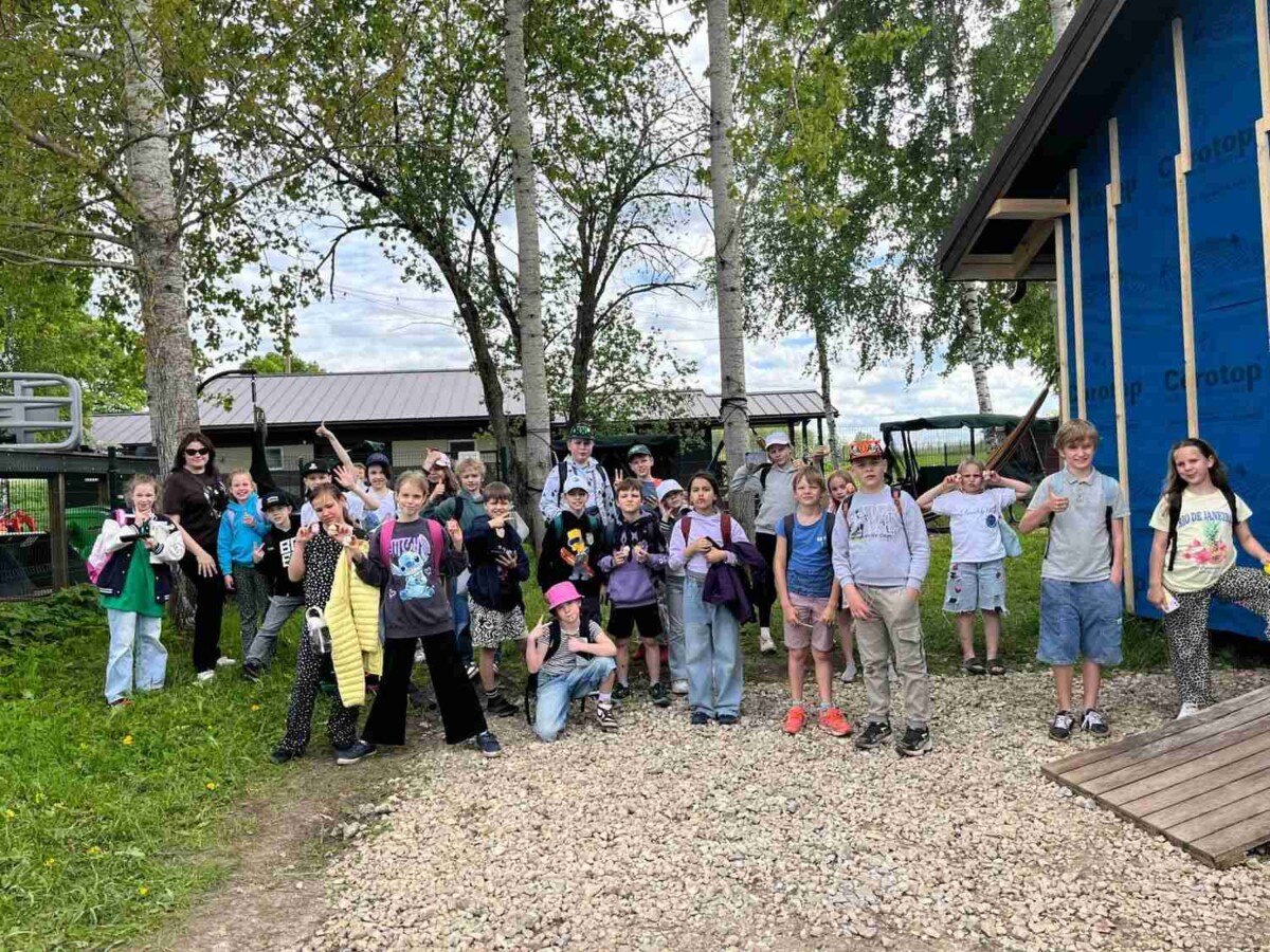 Liela bērnu grupa ar pieaugušajiem pozē fotogrāfijai ārā. Viņi stāv uz grants ceļa, pie zilas ēkas ar apvalku, uz kura redzams uzraksts 