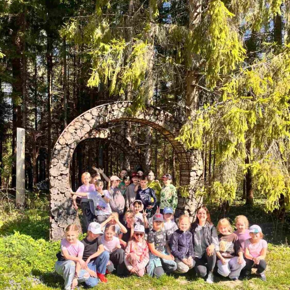 Bērnu un divu pieaugušo grupa pozē fotogrāfijai mežā pie koka baļķīšu arkas. Daži stāv arkā, bet lielākā daļa sēž vai tup priekšā uz zaļas zāles.