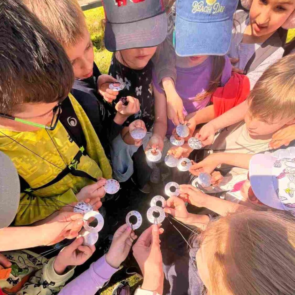 Bērnu grupa ārā tur rokās mazus, apaļus, baltus priekšmetus ar caurumiem; dažiem no tiem ir cipari vai raksti. Viens bērns valkā cepuri ar uzrakstu 