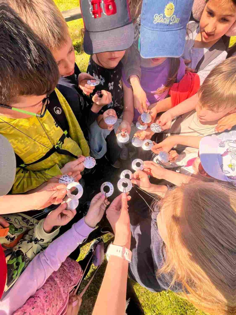 Bērnu grupa ārā tur rokās mazus, apaļus, baltus priekšmetus ar caurumiem; dažiem no tiem ir cipari vai raksti. Viens bērns valkā cepuri ar uzrakstu 