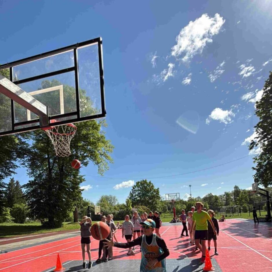 Bērni spēlē basketbolu ārpus telpām saulainā dienā; viens zēns driblē bumbu, kamēr cita bumba lido uz grozu, un citi bērni stāv fonā uz sarkani pelēka laukuma.