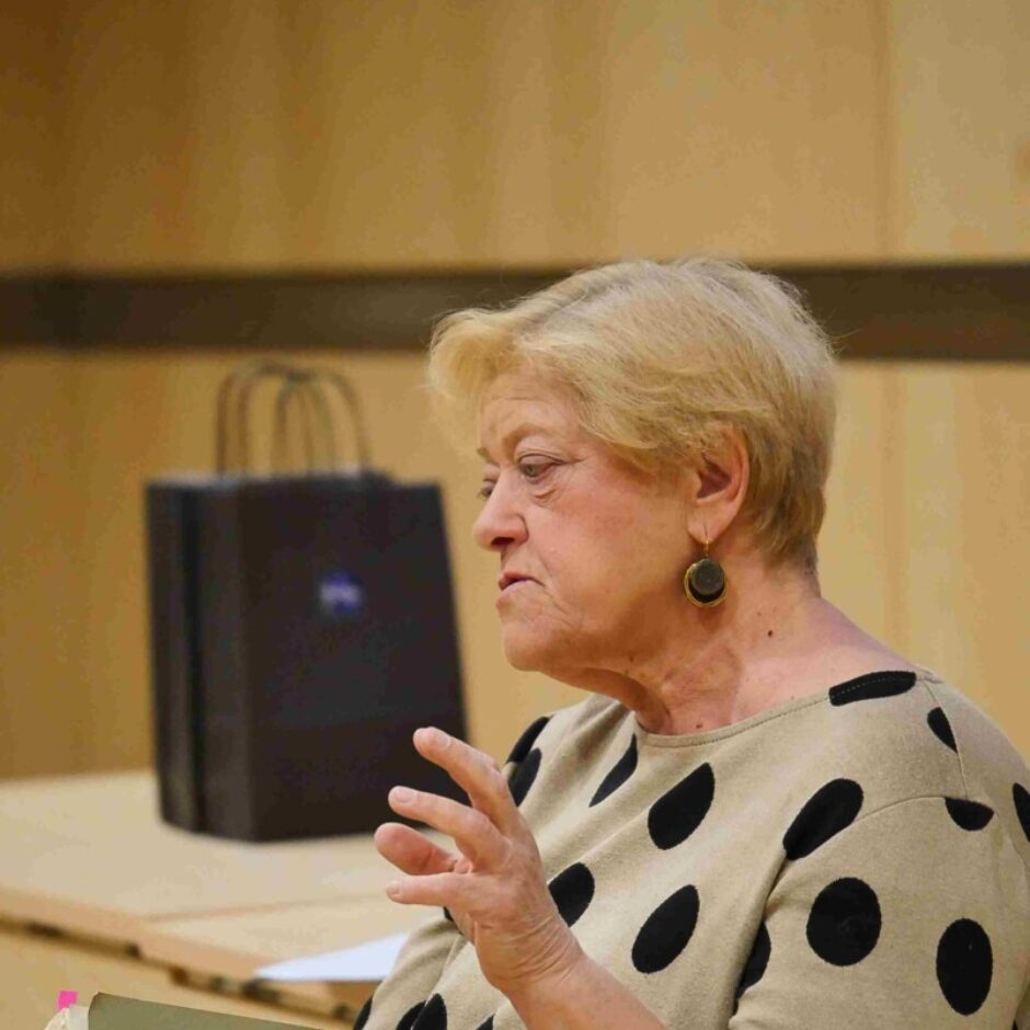 Vecāka sieviete ar blondiem matiem, tērpusies bēšā kleitā ar melniem punktiem, runā un žestikulē ar roku, turot grāmatu ar nosaukumu 
