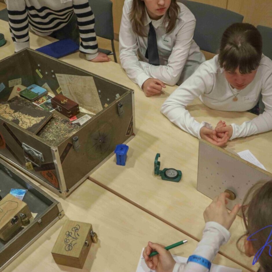 Jauniešu grupa pie galdiem aktīvi risina mīklas, spēlējot spēli ar vairākām kastēm, kartēm un citiem priekšmetiem. Viņi pēta spēles elementus, tostarp mazas koka kastītes, kompasus un papīrus, viena persona nospiež pogu uz baltas plātnes, bet cita tur zīmuli.
