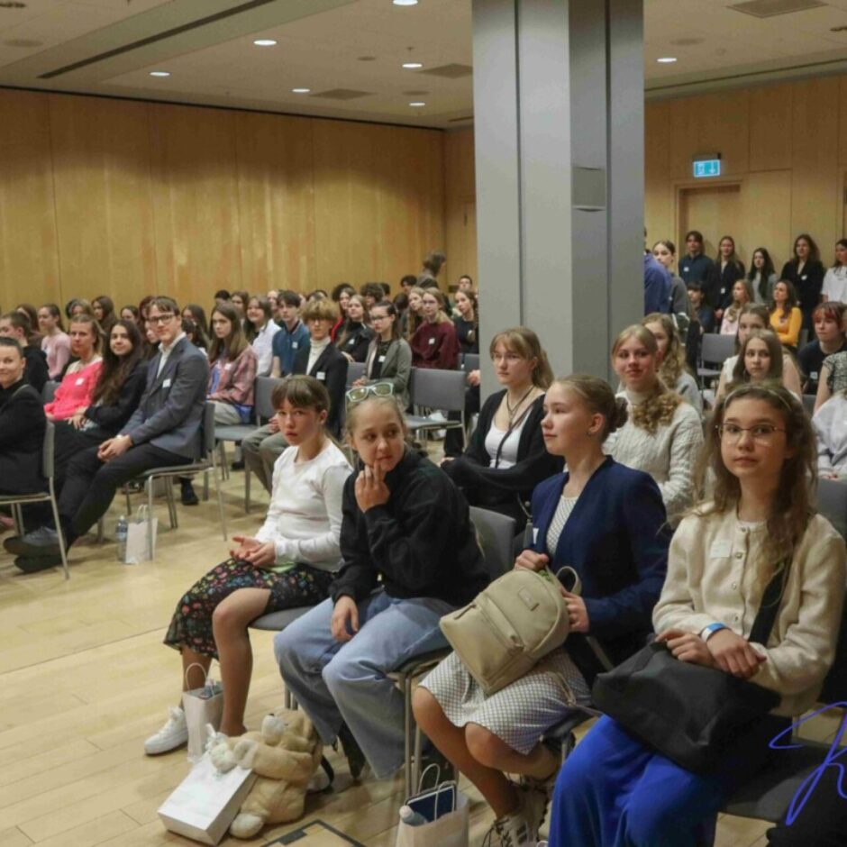 Liela auditorija, galvenokārt jaunas sievietes un pusaudži, sēž krēslos konferenču zālē, skatoties uz priekšu un uzmanīgi vērojot pasākumu. Starp viņiem redzami arī daži pieaugušie.